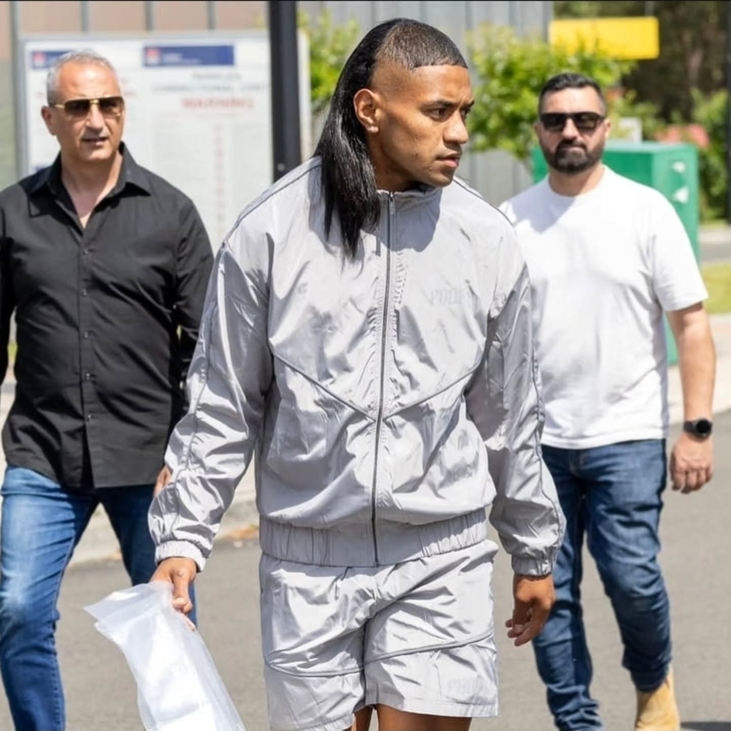 A man with a long slicked-back mullet walks toward the camera in a silver tracksuit, carrying a clear plastic bag, with two men in sunglasses walking behind him.