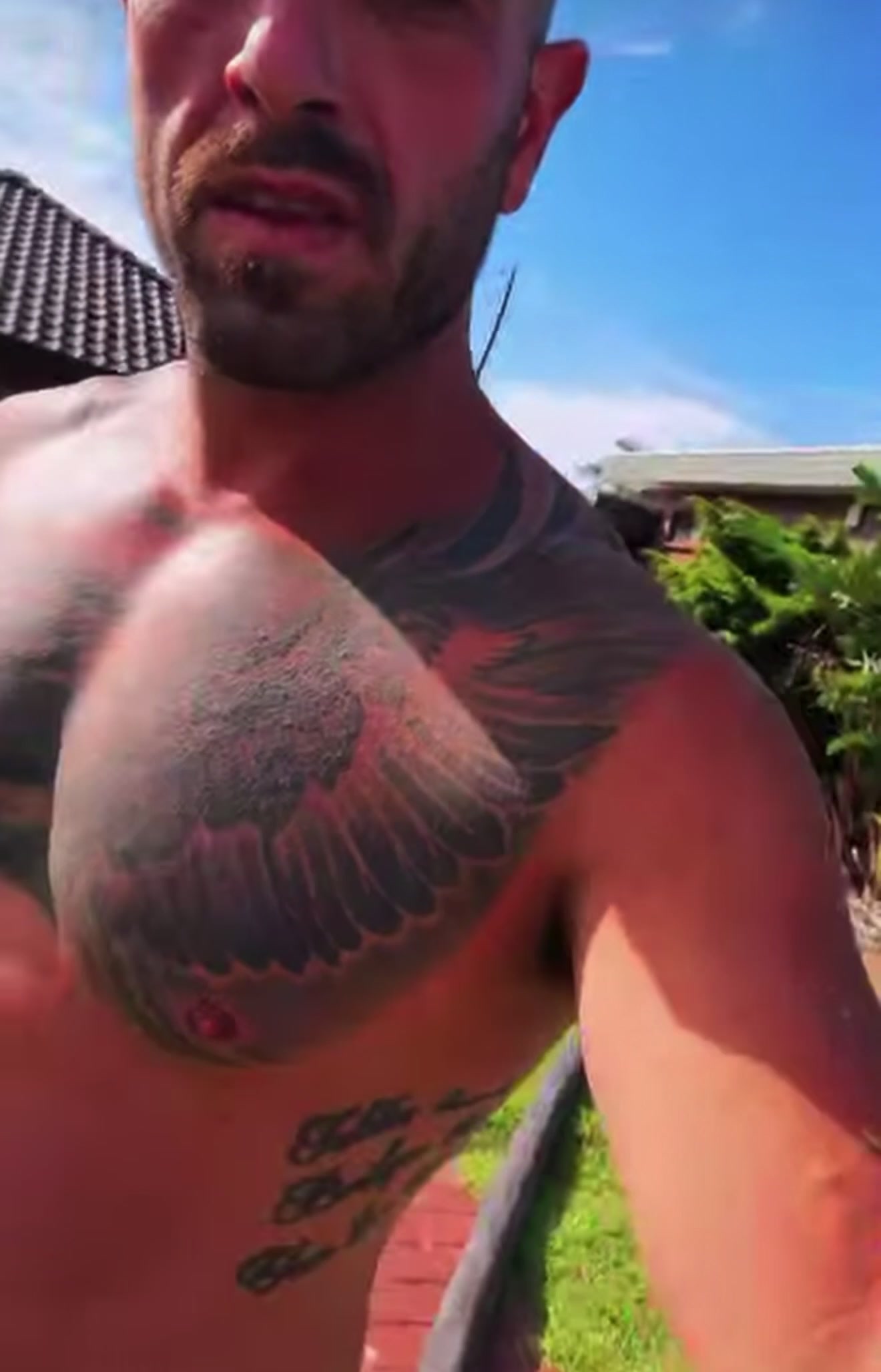 A shirtless, bearded man with a large winged chest tattoo squints into the sun in a sunny backyard with a blue sky behind him.
