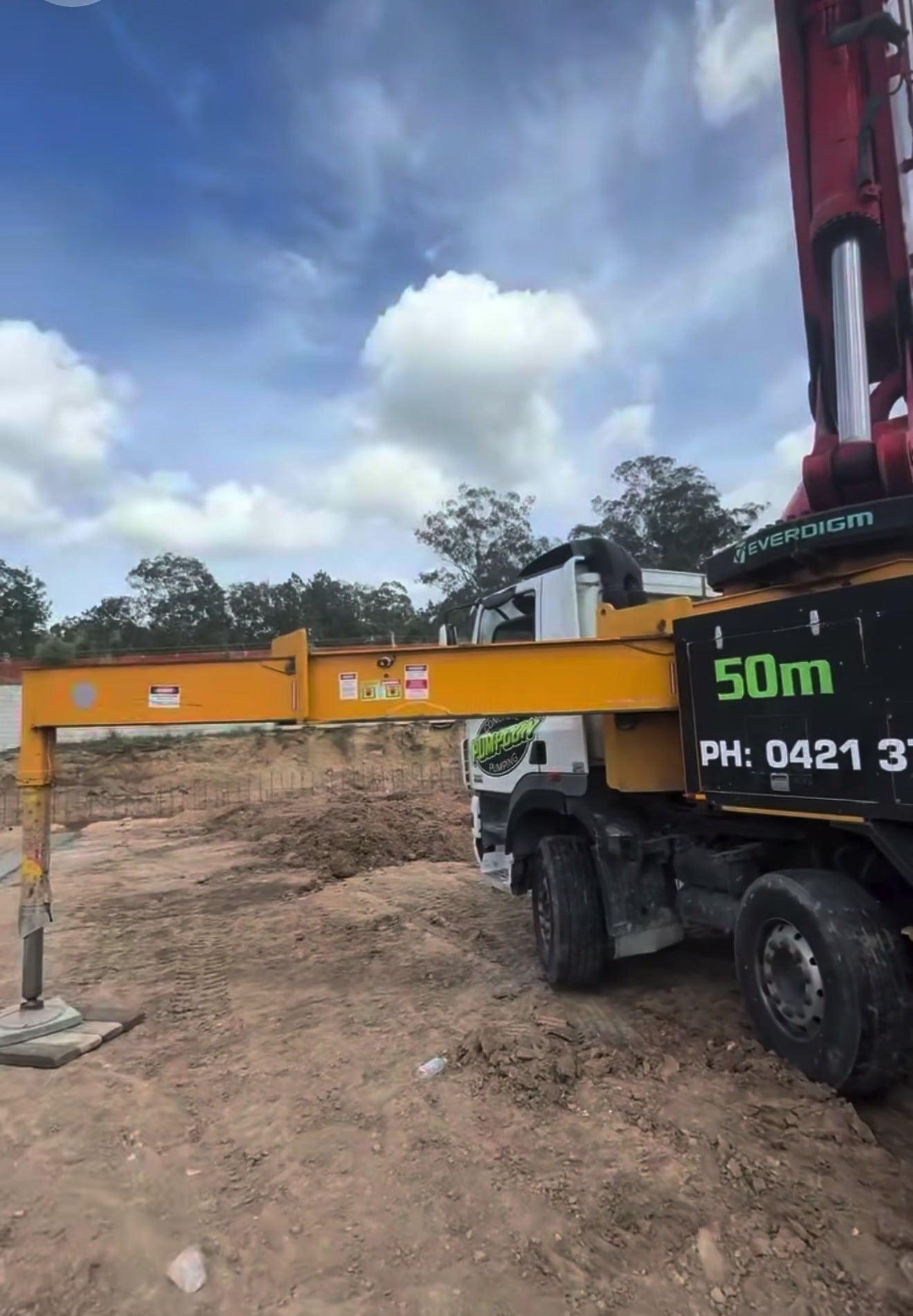 A black, yellow and green 50-metre boom pump truck with its red boom raised and one outrigger extended over dirt under a cloudy blue sky.