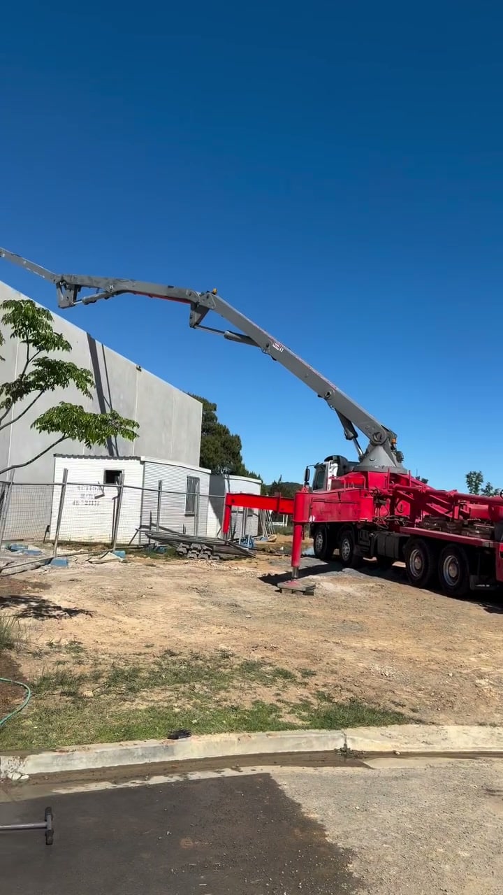 A freshly poured and finished concrete hardstand in front of a row of new industrial units under a clear blue sky, with power trowels and a screed board resting on the slab.