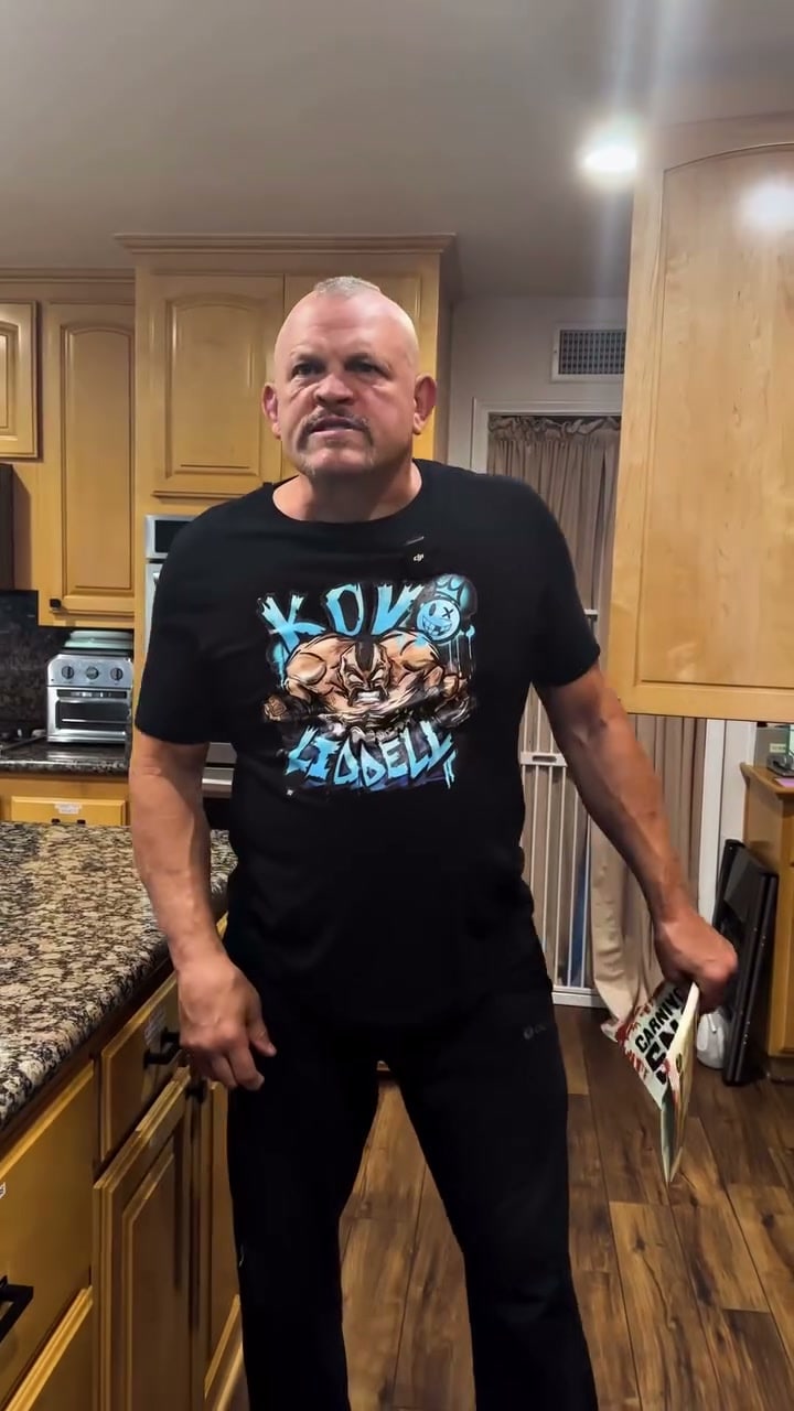 Chuck Liddell scowls in a wooden kitchen wearing a black KO Liddell t-shirt, holding a snack bag in one hand.
