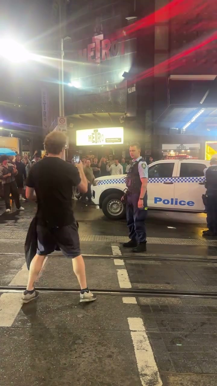 A man in a black tee and shorts holds up a phone on wet tram tracks facing a NSW police officer beside a police ute, with a crowd and the Metro sign behind them at night.