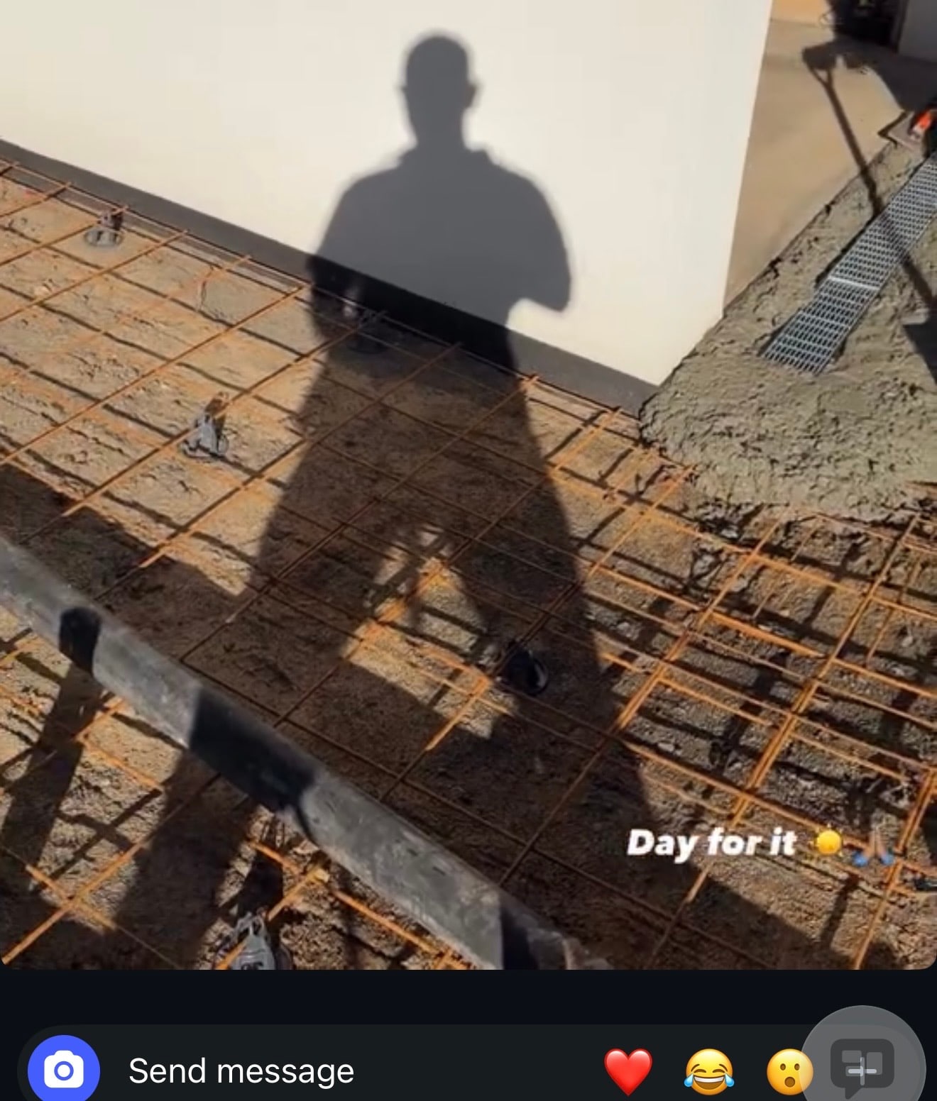 A long human shadow cast across a steel mesh slab and a white wall, with wet concrete and a drain grate at the right and the caption 'Day for it'.