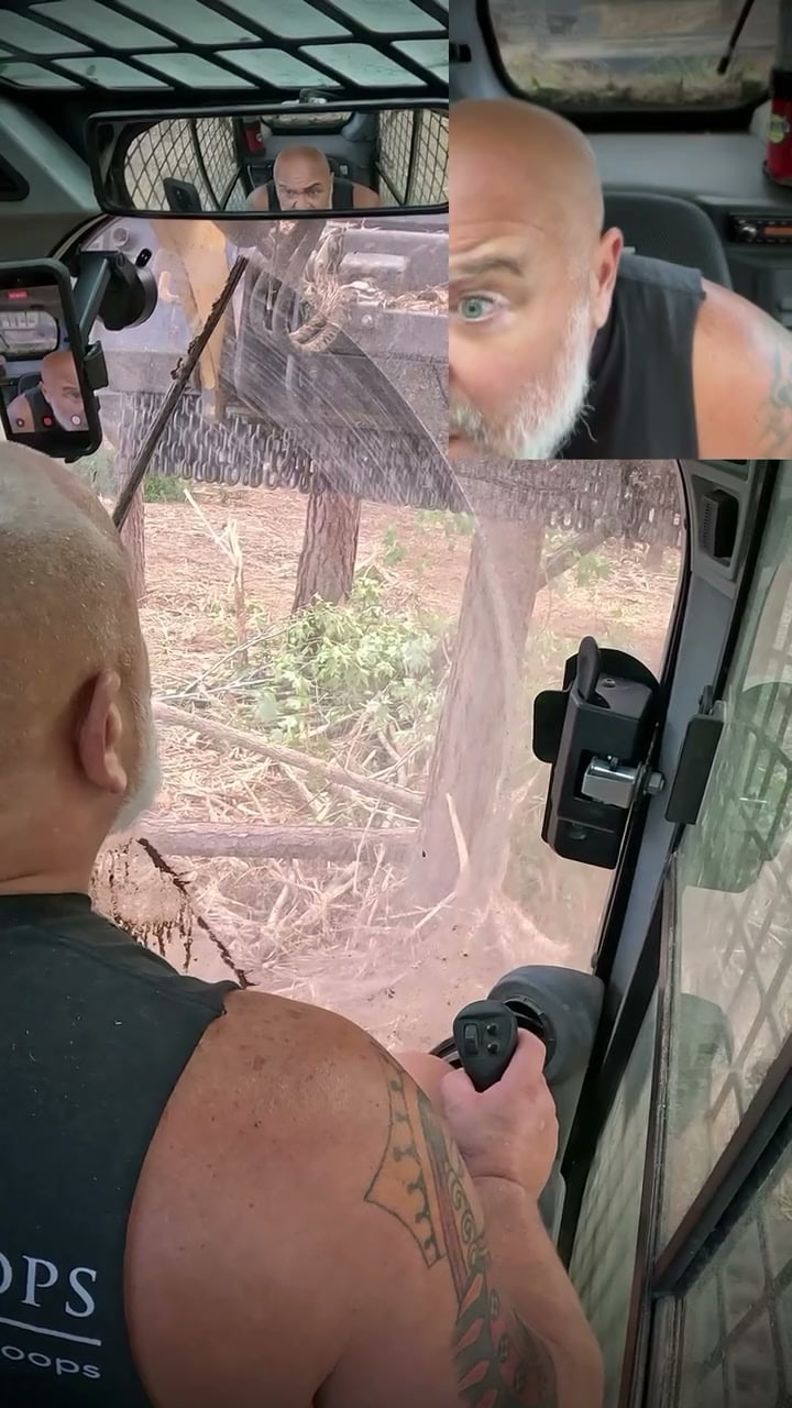 A bald, bearded man in a black sleeveless shirt operates a skid steer with a forestry mulcher while splintered wood sprays across the cab's windscreen, with a close-up of his wide-eyed face in the top corner.