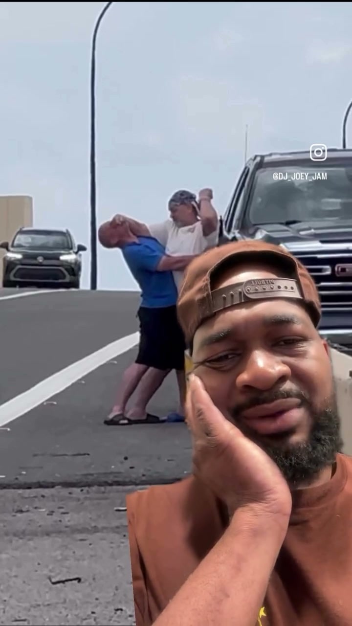 Two men in a headlock-and-slap scuffle on a road beside a pickup, with a bearded man in a backwards cap and orange shirt grimacing in a selfie in the foreground.