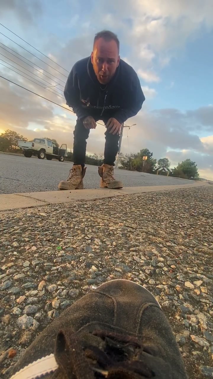 A man in a dark hoodie and work boots leans toward the camera on a footpath, shouting, with a white Hilux ute parked on the road behind him at sunset and the toe of a shoe filling the bottom of the frame.