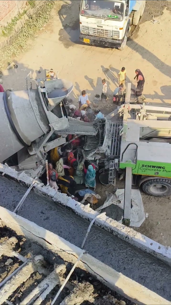 A crowd of people gathered under a tipped concrete mixer chute with buckets while a green Schwing concrete pump stands beside them.
