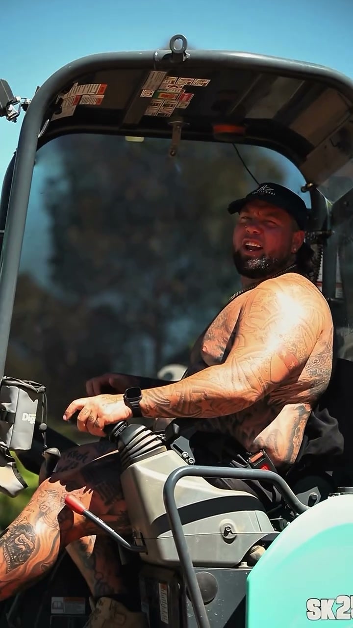 A shirtless crew member laughs knee-deep in a dirt trench beside a shovel, with a mate in a mini excavator and a bearded bloke leaning on the shovel handle.