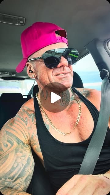 A man in a backwards pink cap, sunglasses, black singlet and gold chain smiles in a vehicle with his seatbelt on.