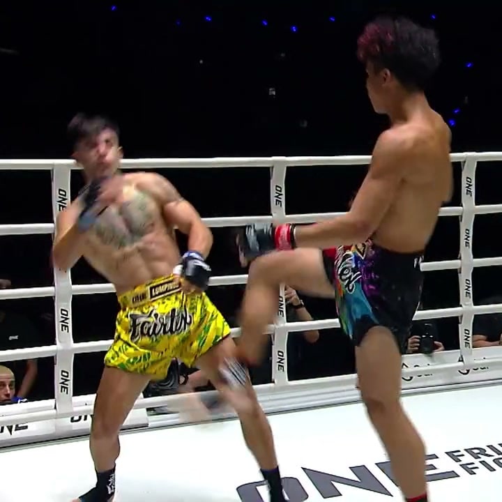 A fighter in rainbow shorts lifts a front kick into the face of an opponent in yellow shorts inside a ONE Championship ring.