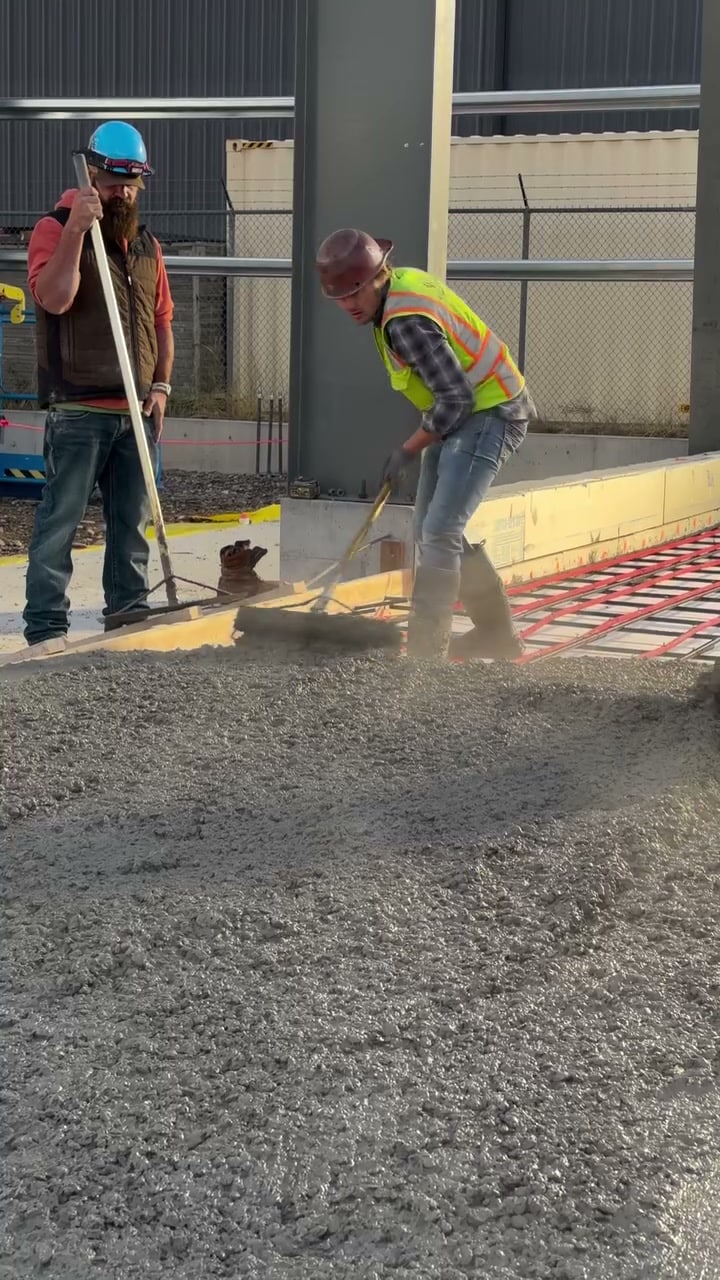 A worker in an orange hi-vis vest, hard hat and muddy boots holds a hose from a red pump sprayer while standing beside a freshly poured, glossy concrete slab.