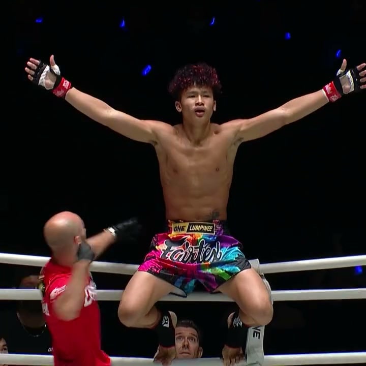 A shirtless fighter in rainbow Fairtex shorts sits on the top ropes with his arms stretched wide while a referee in a red shirt gestures below him.