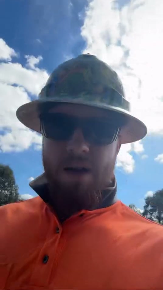 A bearded man in an orange hi-vis shirt, sunglasses and a wide-brim hard hat looks into the camera under a blue sky with clouds.