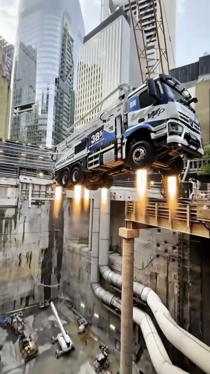 A white and blue concrete boom pump truck hovers above a deep city excavation with jet flames shooting from underneath, surrounded by glass towers.